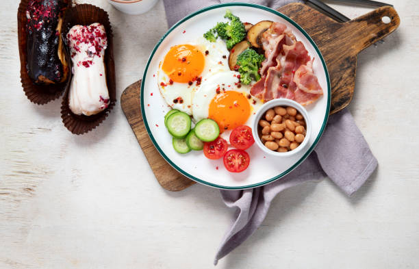 traditional englis breakfast plate with bacon strips, sunny side up eggs, vegetables and cake on light background - englis imagens e fotografias de stock