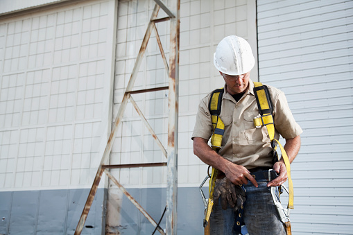 Worker (30s) with work gloves, hard hat, safety glasses, putting on harness.