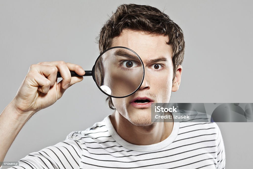 Man with magnifying glass Curious man looking through magnifying glass at the camera. Studio shot, grey background. Magnifying Glass Stock Photo