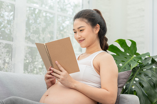 Happy Pregnant Woman sitting on couch holding book and reading for prepare to take care baby,Pregnancy of young woman enjoying with future life relax at home,Motherhood and Pregnant Concept,Soft focus