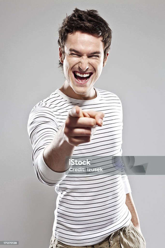 You! Portrait of laughing young man pointing at the camera. Studio shot, grey background. Front View Stock Photo