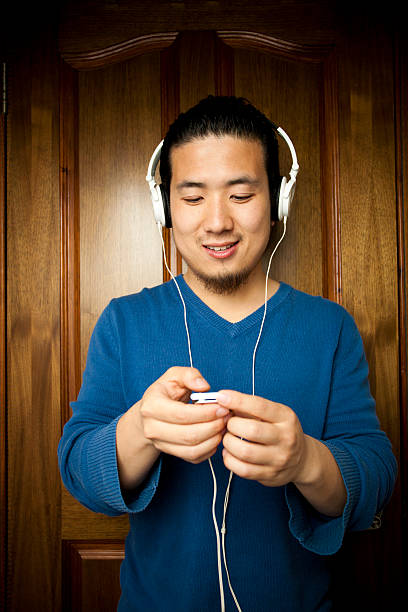 Brightly lit isolated image on white background of an Asian young woman with black and long hair in white spaghetti strap top. She is lying or sleeping on white bed and pillow with closed eyes and listening songs with earphones on through a smart phone. Horizontal composition with copy space at aperture f/2.