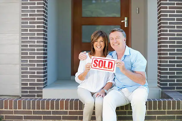 Happy mature couple holding sold sign in front of a new house