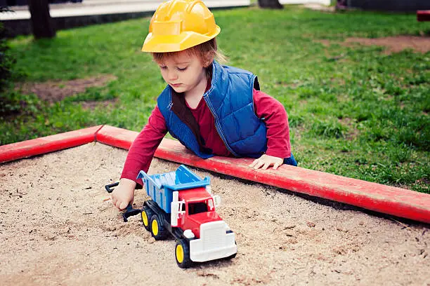 "Two years old boy, wearing yellow construction helmet and blue waistcoat, crouching near sandbox and playing with toy truck.Similar:"