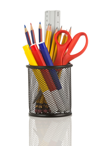 A gray wired pencil holder full of some office or school supplies against a white background.  There are multicolored items in the pencil holder like a ruler, scissors, yellow pencils, and markers. The reflection of the gray wires can be seen faintly in the gleaming white surface in the foreground. DSRL studio picture taken with Canon EOS 5D Mk II.