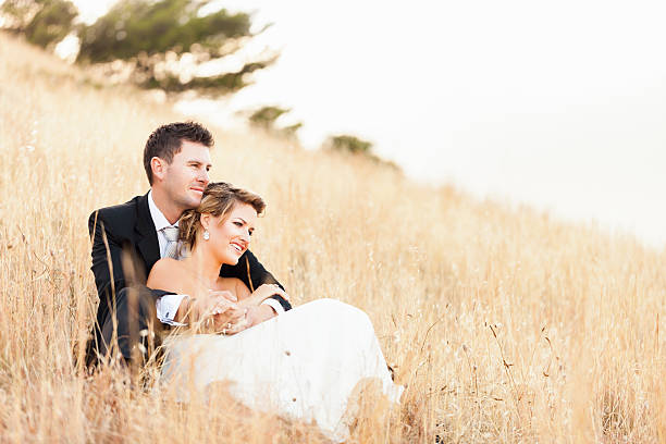 Happy newlyweds sitting in dry grass, looking into copy space
