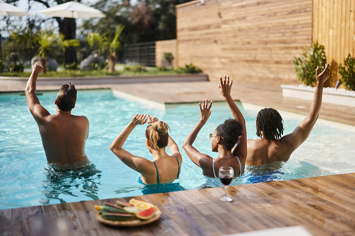 Rear view of carefree diverse couples having fun while dancing with their arms raised during summer party in the pool.
