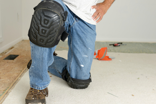 A man's legs as he kneels on a carpet he is removing from a room.