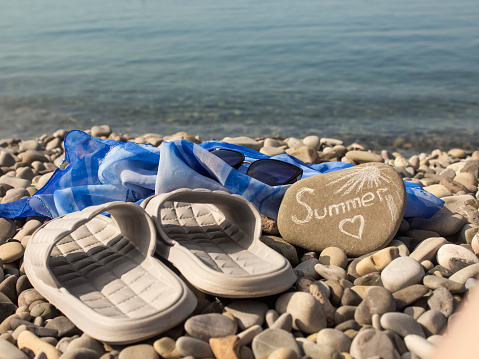 gray flip-flops and a blue pareo, sunglasses and a stone with the inscription Summer on a pebble beach.