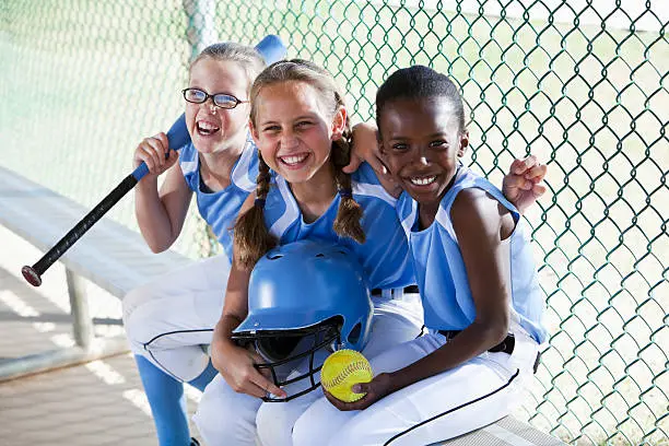 Multi-ethnic girls (9 years) on softball team at the ball park, sitting in dugout.