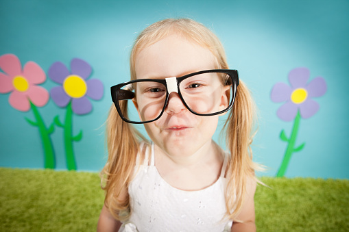 Color image of a nerdy, little blond-haired girl with big glasses making a face, in a whimsical, outdoor world.