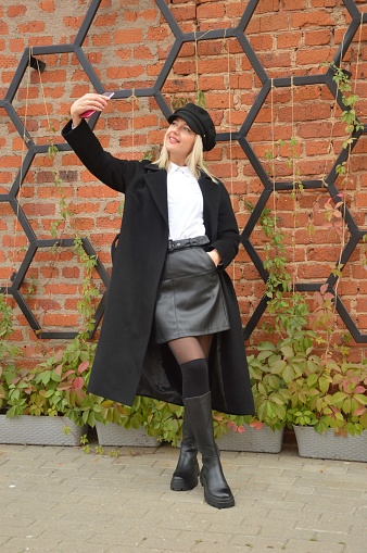 Young woman, blonde girl taking selfie with smartphone in black coat and stylish cap on city street