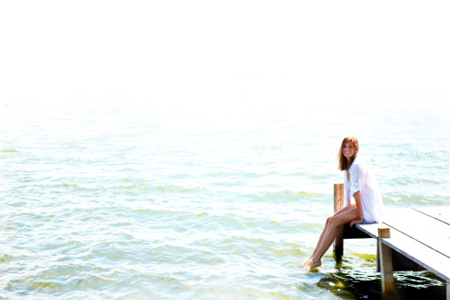 A beautiful young woman sitting on the edge of a jetty with her feet in the water - copyspace