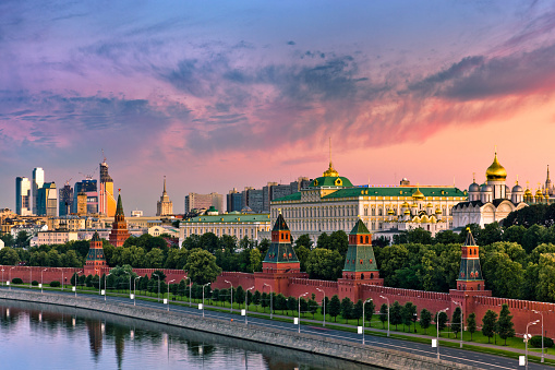 View of the Moskva River along the Kremlin wall in Moscow, Russia, with water and a walkway in the foreground and a beautiful sunset sky in the background.  The image shows the Grand Kremlin Palace and the cathedrals of the Moscow Kremlin.  The view is from the top of the Hotel Baltschug Kempinski.