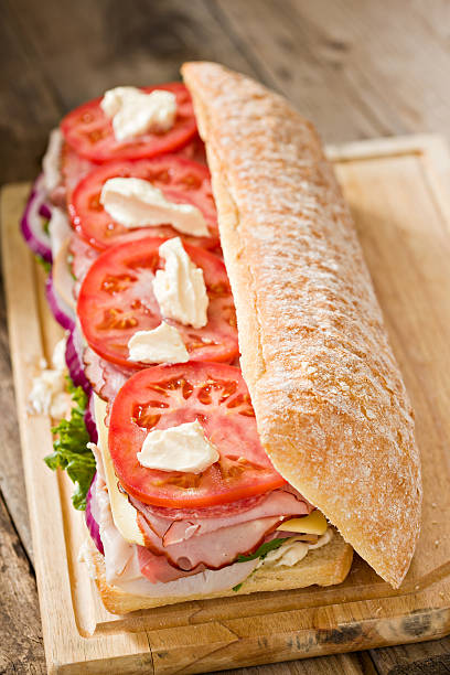 Giant Dagwood Sandwich "A high angle close up of a giant dagwood sandwich consisting of a large ciabatta roll, mayonnaise, lettuce,red onions smoked turkey,roast beef, jarlsberg cheese, green bell peppers, bavarian ham, italian dry salami, and tomatoes. Shot against an old wooden background." dagwood stock pictures, royalty-free photos & images