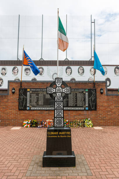 Visitors stand before the Clonard Martyrs Memorial in Belfast, Northern Ireland. The large cross and commemorative plaques honor those lost during the Troubles, with mural portraits displayed above.