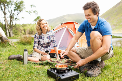 Happy mid adult woman holding cup while man cooking food on a portable camping stove. Horizontal shot.