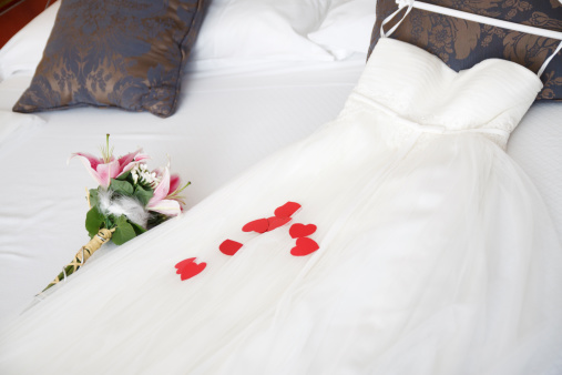 A beautiful white wedding dress and a bouquet  with red hearts on it laying down on a bed. The dress is waiting for the bride to put it on.