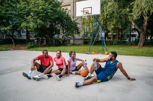Young black family sitting on the basketball court outdoors,chatting and drinking water after a basketball game.