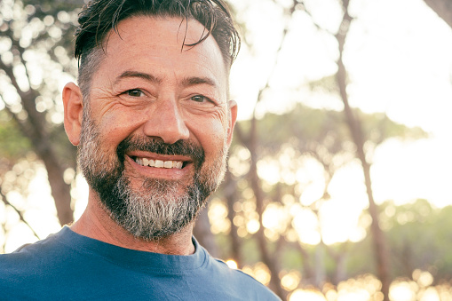 Cheerful outdoor portrait of happy adult mature man smiling at the camera with trees and sunset light in background. Beard and serene expression. One male people enjoy lifestyle outdoors alone