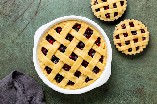 Cooking mixed berry crust pie or tart with lattice, mini tarts ready to put in the oven for baking. Raw dough or shortcrust pastry, fresh berries. Green table surface, directly above.