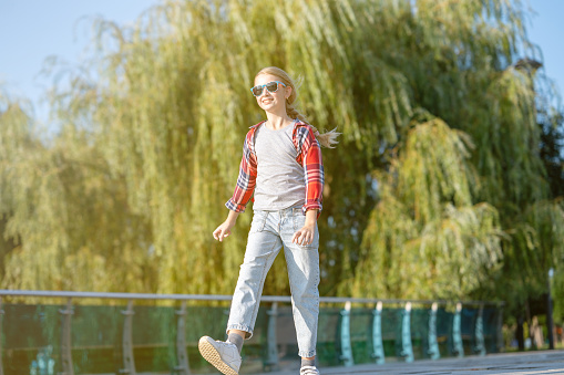 A girl in a plaid shirt jumps and runs along a path in a city park on a sunny summer day. Teenage holiday concept.