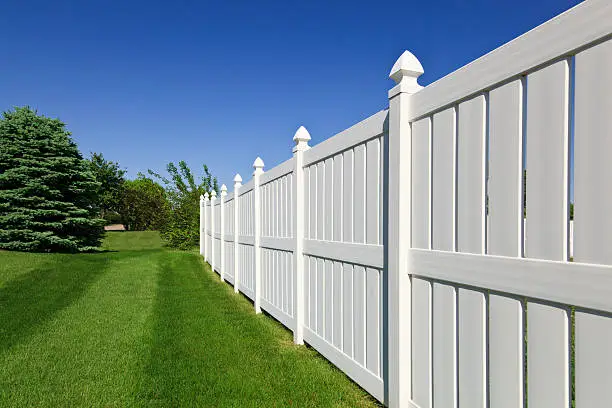 New and contemporary white vinyl fence running across a nicely landscaped backyard with lawn and blue sky in the background.