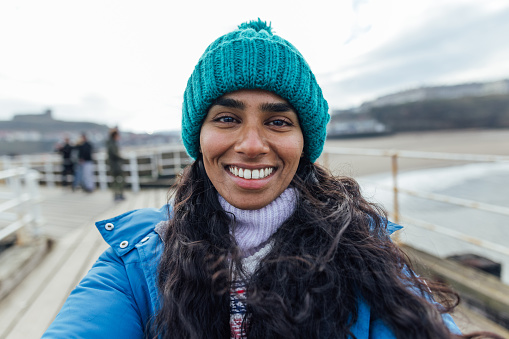 A selfie shot of a mid-adult woman wearing warm casual clothing and accessories. She is enjoying a day out in the seaside town of Whitby in February. She is looking and smiling at the camera.