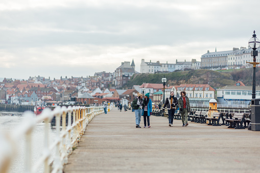 A wide shot of two heterosexual couples wearing warm casual clothing and accessories. They are enjoying a day out in the seaside town of Whitby in February. They are walking along the pier, talking and laughing.\n\nVideo is also available for this scenario.