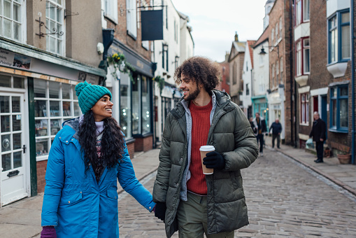 A medium shot of a heterosexual couple wearing warm casual clothing and accessories. They are enjoying a day out in the seaside town of Whitby in February. They are walking down a cobblestone street, holding hands and looking at each other.