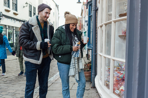 A medium shot of a heterosexual couple wearing warm casual clothing and accessories. They are enjoying a day out in the seaside town of Whitby in February. They are standing on a cobblestone street with takeaway coffees and snacks in hand and looking in a shop window.

Video is also available for this scenario.