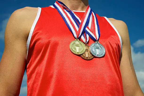 Athlete in red athletic shirt stands with gold, silver, and bronze medals in front of bright blue sky