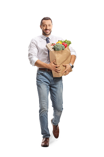 Full length portrait of a smiling professional man walking and carrying a bag with fruits and vegetables isolated on white background
