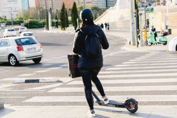 giovane donna con casco e scooter elettrico attraversa un marciapiede in un'area urbana della città, uno spostamento sostenibile e non inquinante. - nonpolluting foto e immagini stock