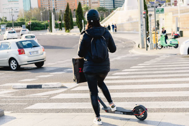 młoda kobieta z hełmem i skuterem elektrycznym przechodzi przez przejście dla pieszych w miejskiej części miasta, zrównoważone i niezanieczyszczające wysiedlenie. - nonpolluting zdjęcia i obrazy z banku zdjęć