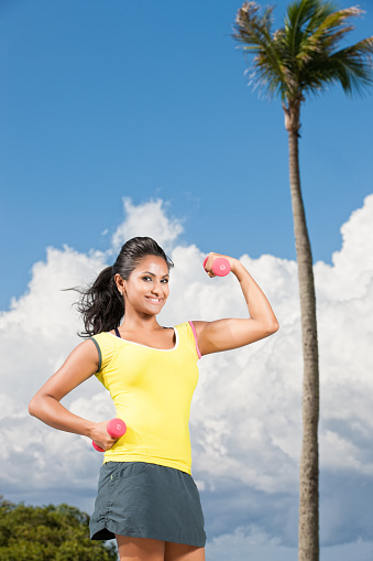 A beautiful ethnic woman holding exercise weights outside by a palm tree, flexes her muscles, proud of her fitness.