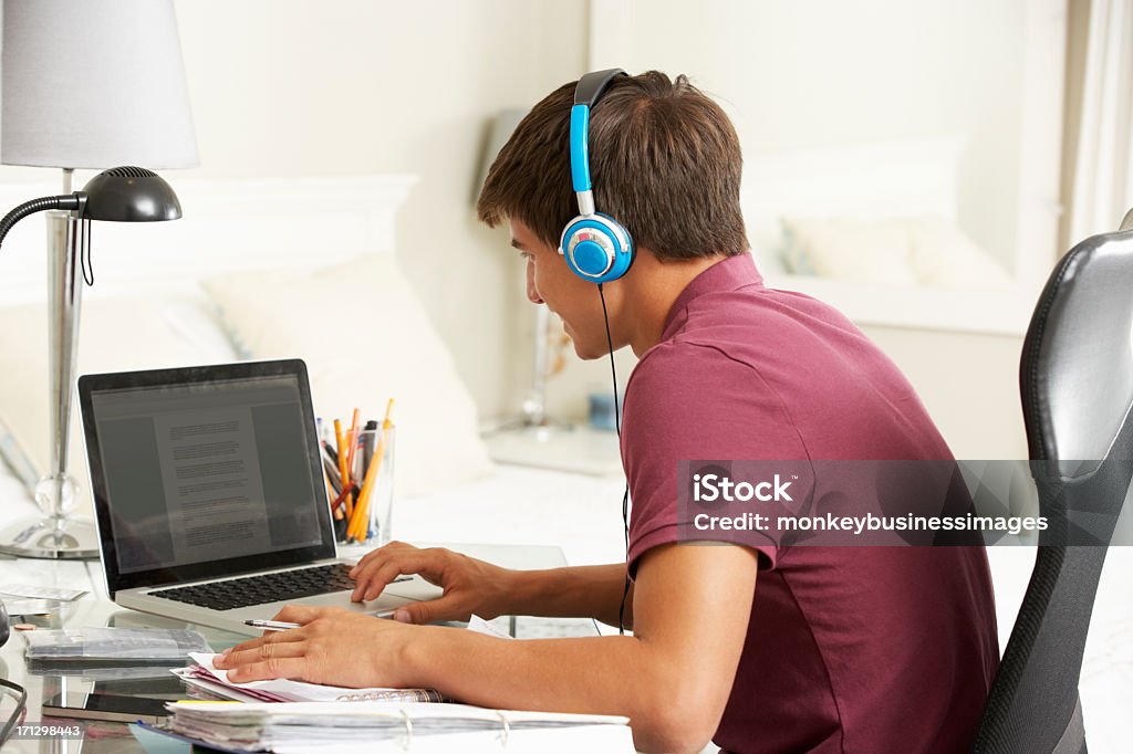 Teenage Boy Studying At Desk In Bedroom Wearing Headphones Teenage Boy Studying At Desk In Bedroom Wearing Headphones Smiling Looking At Computer Bedroom Stock Photo