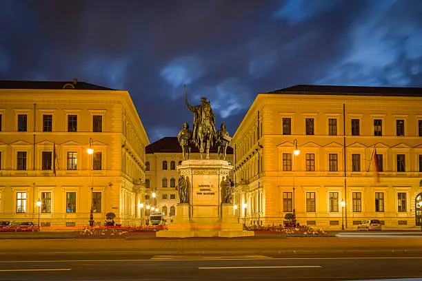 The square Odeonsplatz in Munich, Germany, with the statue of King Ludwig I., King of Bavaria. Night shot with dark blue sky - blue hour. Long exposure - a trace of a red backlight of a car.