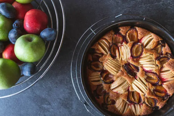 Delicious and juicy autumn winter cake with apples and plums. Served with a bowl of fresh fruits in a baking pan on dark background from above.