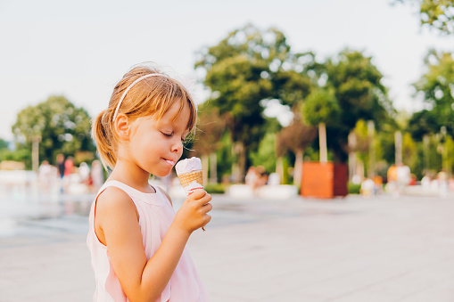 Little preschool girl eating ice cream in waffle cone on sunny summer day. Happy toddler child eat icecream dessert. Sweet food on hot warm summertime days. Bright light, colorful ice-cream.