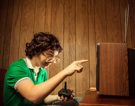 A curly haired retro styled 1980's teenager turns on his television to play Video console games, a goofy grin on his face. Vintage wood paneling on the walls.  Horizontal with copy space.