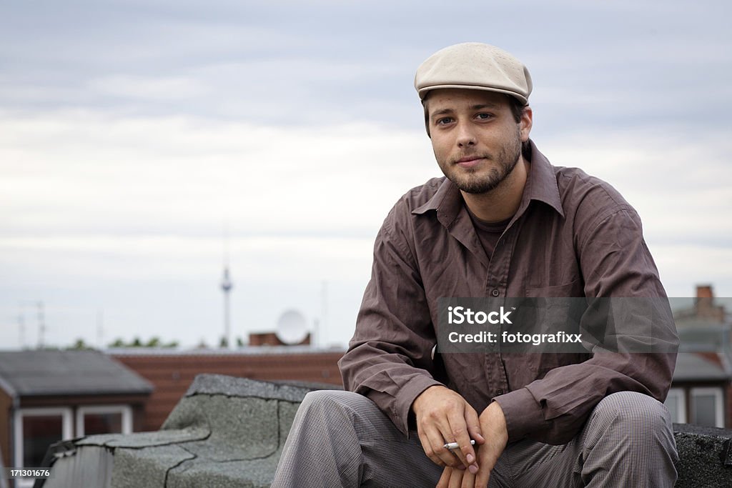portrait of a handsome smoking man on a roof portrait of a handsome young man sitting on a roof in Berlin, smoking a cigarette, tv tower in the background Flat Cap Stock Photo