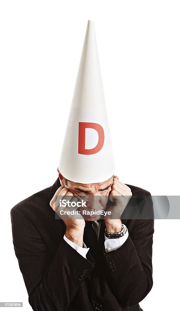 Businessman in dunce cap looks embarassed grumpy and resentful "Caught out in some misdemeanor, a businessman wearing a dunce's cap has his head in his hands, looking down grumpy, rebellious and embarrased. Isolated on white." 30-39 Years Stock Photo
