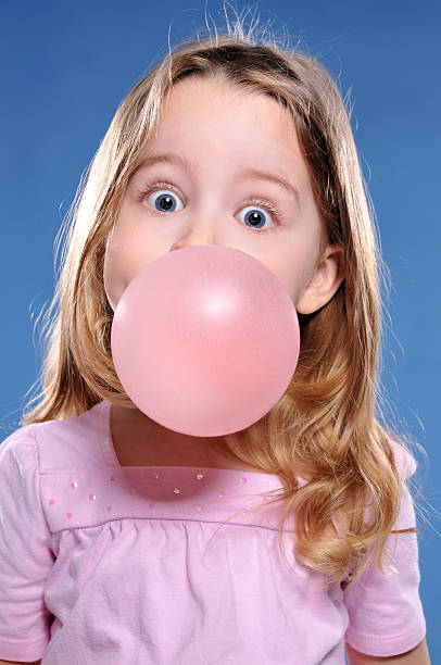 vertical image of an adorable 5 year old girl dressed in pink with a dark blue background. she is blowing a big bubble with her gum and it's about to pop. great expression. rim lights brighten her long blonde hair.