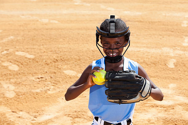 Softball player African American girl (9 years) playing softball, wearing face mask (standard safety gear required by many leagues). softball pitcher stock pictures, royalty-free photos & images