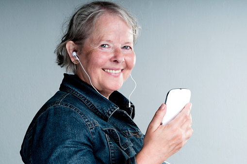 Happy senior woman is looking at the camera. She is holding a smart phone and she smiles. Earbuds are connected to the phone. Maybe she is listening to music, an audio book or a podcast. 