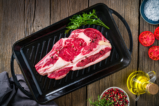 Beef rib steak raw on a cast iron grill background uncooked with herbs olive oil, tomato, pepper ingredients on rustic dark wood table, top view flatlay