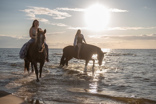 Two women in equestrian attire gallop through the surf on the beach, with the waves of the ocean providing a picturesque backdrop