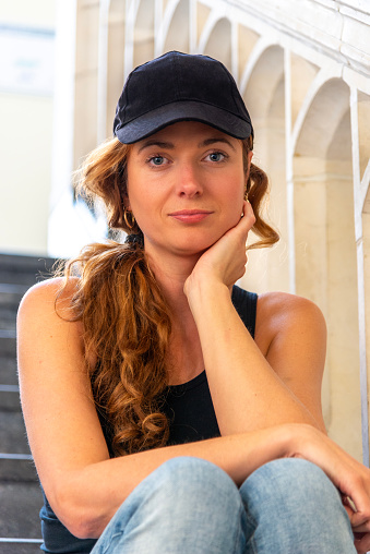 Portrait of a beautiful hipster woman with a black cap, situated on a staircase, looking at camera.