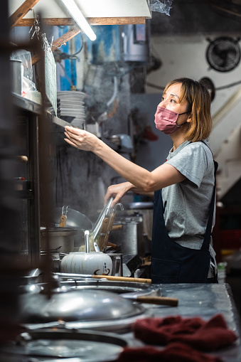 Amid the hustle and bustle of her noodle shop, the proprietress takes a moment to glance up and confirm an order before resuming her meticulous noodle-cooking process, ensuring each dish meets her high standards.